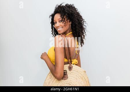 Back view of cheerful ethnic Woman with wicker bag looking at camera over shoulder on light background Stock Photo