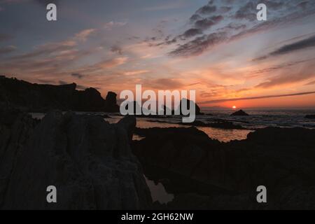Amazing peaceful scenery of sunset over rippled wavy sea with rocks under colorful cloudy sky in summer evening in Liencres Cantabria Spain Stock Photo
