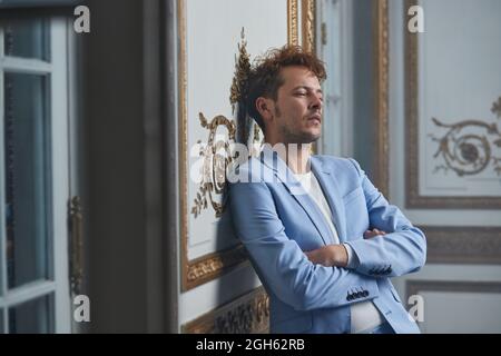 Thoughtful male in suit standing with crossed arms and leaning on wall in posh room Stock Photo