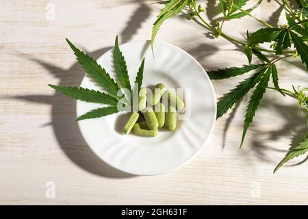 Prescription herbal pills of cannabis sativa weed and green marijuana leaf on a white saucer near fresh hemp stem with green leaves over wooden table. Stock Photo