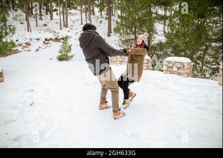 Delighted couple in warm clothes holding hands and spinning around in snowy winter woods while having fun Stock Photo