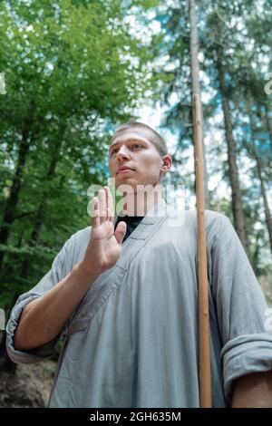 Low angle of man with stick holding hand near chest while practicing kung fu in woodland Stock Photo