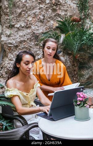 Multiracial female freelancers sitting at table in tropical cafe and working on project while typing on keyboard dock of tablet Stock Photo
