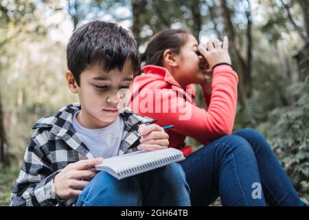 Ethnic boy writing in notepad against sister looking through binoculars while sitting on land in summer woods Stock Photo