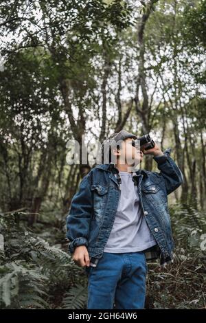 Ethnic child in denim jacket looking through binoculars while exploring woods with overgrown trees and lush plants Stock Photo