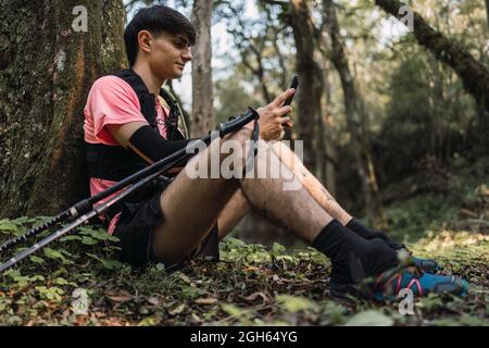 Side view of content male hiker with backpack sitting in woods and browsing mobile phone while traveling Stock Photo