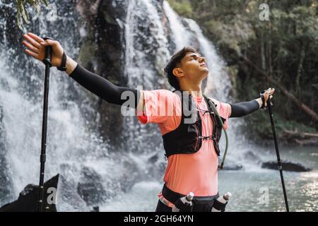 Dreamy male hiker with trekking poles in outstretched hands standing with closed eyes near waterfall in forest and enjoying freedom Stock Photo