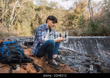 Thirsty male hiker with backpack drinking water from cup with straw while sitting on rock near waterfall in forest and look-in away during break Stock Photo