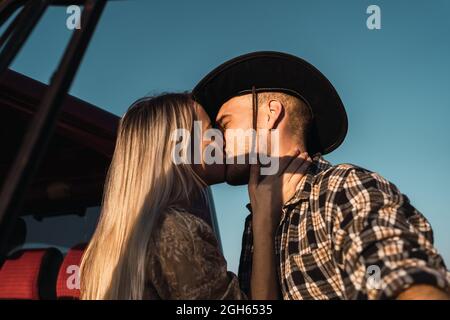 From below side view of young loving Woman kissing man in cowboy hat tenderly near car on background of blue sky in evening Stock Photo