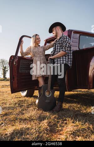 boyfriend in cowboy hat with acoustic guitar while standing with girlfriend in red retro pickup car parked on sandy road in countryside Stock Photo