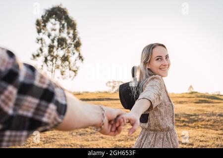 Cheerful Woman holding hand of crop man while standing in meadow in summer evening and showing follow me concept Stock Photo