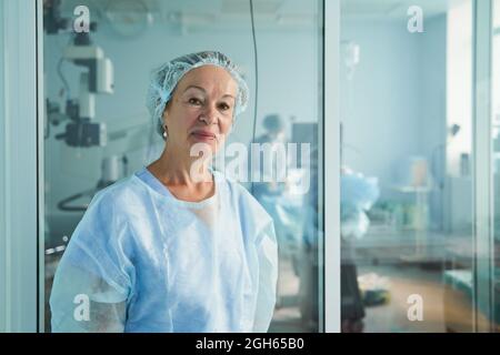 Friendly middle aged female doctor in surgical uniform looking at camera against unrecognizable colleague in hospital Stock Photo