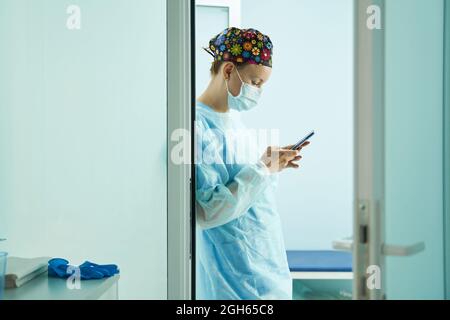 Side view of female doctor in medical uniform and sterile mask text messaging on cellphone in clinic Stock Photo