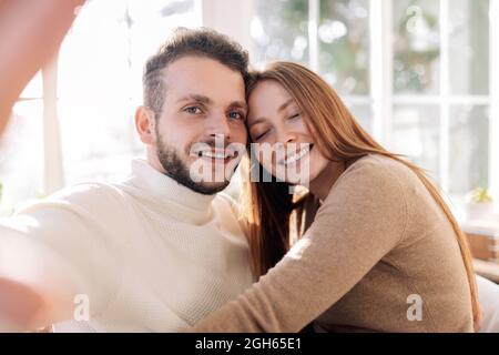 Cheerful bearded man with sincere female beloved taking self portrait while looking at camera at home Stock Photo