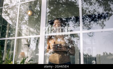 Through window view of contemplative young female with cup of hot beverage looking away against window in house Stock Photo