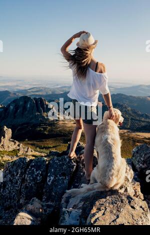 Back view of young Woman with Golden Retriever dog standing on rock and admiring breathtaking mountains during summer trip Stock Photo