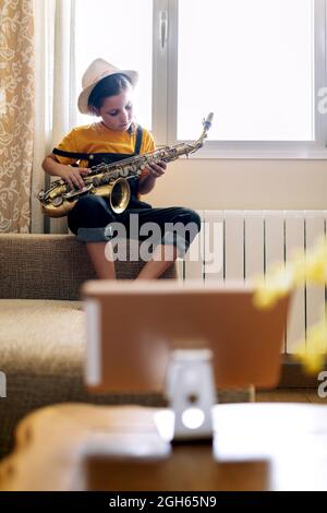Child playing saxophone while sitting on couch recording video on mobile phone against window in room Stock Photo