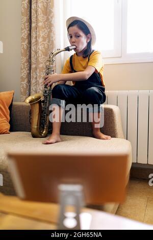 Child with saxophone sitting on couch while recording video on mobile phone against window in room Stock Photo