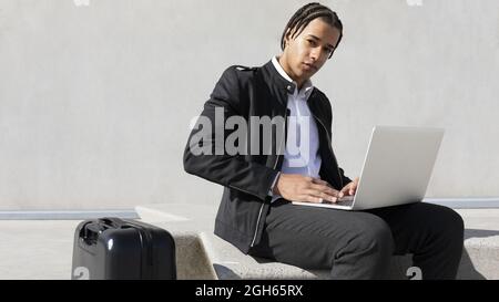 Young ethnic male freelancer with braided hairstyle sitting on bench and typing on laptop keyboard while working on project remotely on urban street Stock Photo