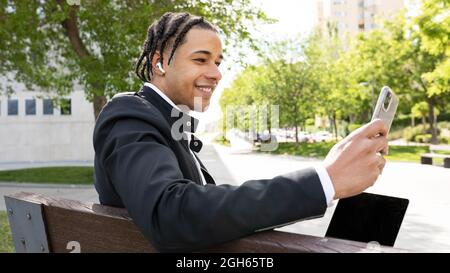 Side view of smiling young ethnic businessman with braids making video call using smartphone and TWS earbuds while sitting on bench with laptop on urb Stock Photo
