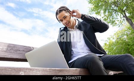 From below of happy ethnic male freelancer with braided hairstyle sitting on bench and typing on laptop keyboard while working on project remotely on Stock Photo