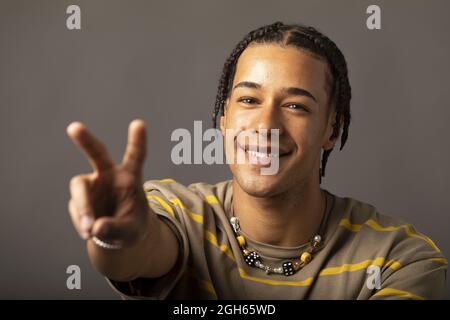 Happy young African American hipster guy with braids wearing striped shirt and necklace looking at camera and showing two fingers sign against grey ba Stock Photo