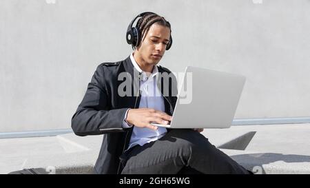 Young focused African American male manager with braids in formal suit and wireless headphones while working on laptop on urban street Stock Photo