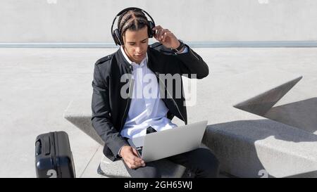 Young focused African American male manager with braids in formal suit and wireless headphones while working on laptop on urban street Stock Photo