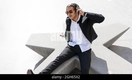 High angle of young ethnic male with braided hair dressed in stylish suit and sunglasses enjoying music through wireless headphones while resting on s Stock Photo