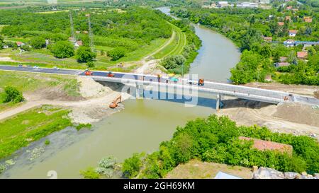 Above view, sideways, on workers and machinery for laying asphalt, spreading layer of hot tarmac over unfinished bridge, road over river is under cons Stock Photo