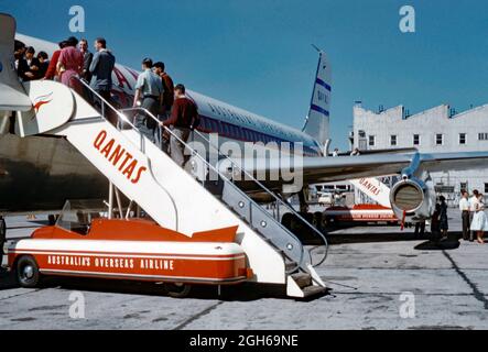 Passengers getting on a Qantas Boeing 707 aeroplane in 1960 by means of a mobile boarding stairs vehicle. Qantas Airways Limited is the flag carrier of Australia and its largest airline. It is the world's third-oldest airline still in operation, having been founded in November 1920. The Qantas kangaroo logo is on the staircase and the plane’s tail. Note the streamlined art-deco curves of the boarding vehicle. This image is from an old amateur Kodak colour transparency – a vintage 1960s photograph. Stock Photo
