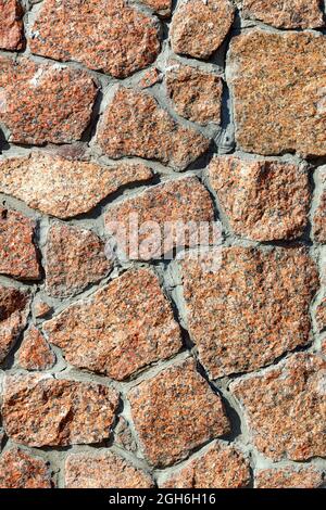 Texture of a red granite wall embedded in a wall in warm sunlight, close-up, high resolution. Vertical image. Stock Photo