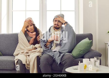 Young unhappy couple in warm clothes and blankets sitting on sofa, touching foreheads and feeling ill Stock Photo