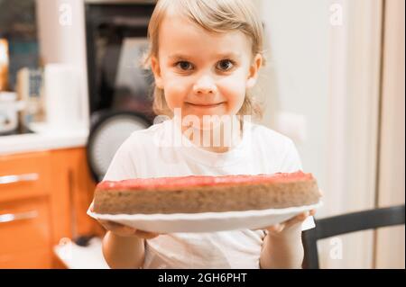 portrait of a cute little happy candid caucasian five year old kid boy in white t-shirt holds a homemade baked cake or pie in his hands at home Stock Photo