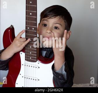 Little hispanic boy shows Rock and Roll Hand Signs and holding an electric red guitar isolated on white background. Sign of the horns Stock Photo