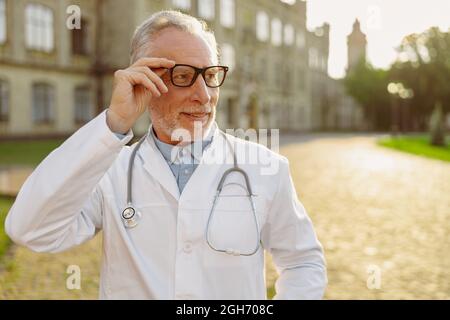 Portrait of serious senior male doctor in lab coat adjusting his glasses, looking aside, standing outdoors near clinic Stock Photo