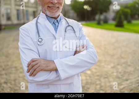 Cropped shot of smiling senior male doctor in lab coat standing with arms crossed outdoors near clinic Stock Photo