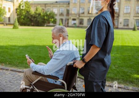 Senior man, recovering patient in wheelchair making video call using smartphone while walking with nurse wearing face shield and mask Stock Photo