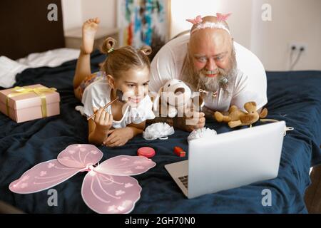 Little girl and father with cosmetic brushes watch beauty blogger video on bed at home Stock Photo