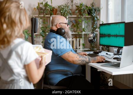 Plus size man with headphones works on computer while girl holds gift box in room Stock Photo