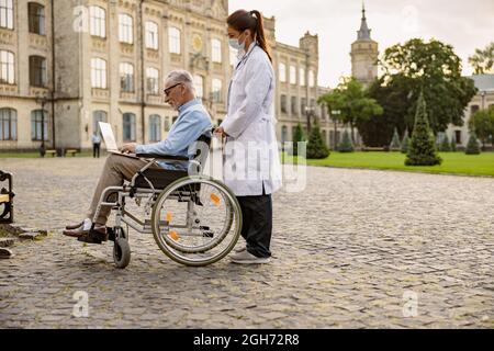 Full length shot of young female doctor taking care of senior recovering patient in wheelchair using laptop, walking outdoors in the park near clinic Stock Photo