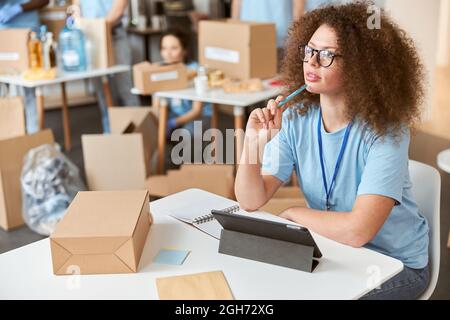 Thoughtful young woman, volunteer in blue uniform looking away, using tablet pc while sitting and working on donation project. Team sorting, packing Stock Photo