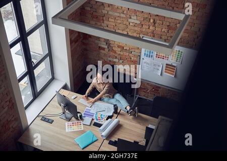 Top view of relaxed woman designer typing while working on new interior design project, sitting at the table in modern office Stock Photo