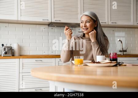 Mature grey woman smiling while having breakfast in kitchen at home Stock Photo