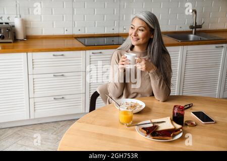 Mature grey woman smiling while having breakfast in kitchen at home Stock Photo