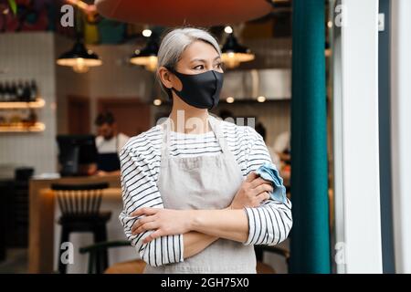 Mature waitress woman wearing face mask standing in cafe indoors Stock Photo