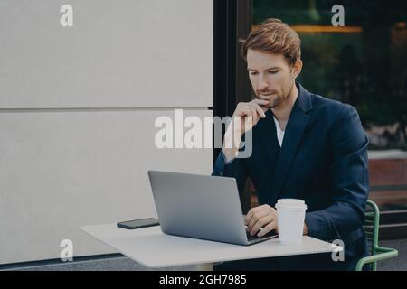 Handsome pensive young man browsing web on laptop while working remotely in sidewalk cafe Stock Photo