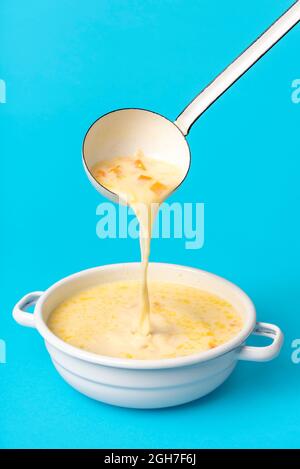 Delicious greek soup in white enamel bowl isolated on a blue background. Pouring soup from an enamel ladle into the bowl. Stock Photo