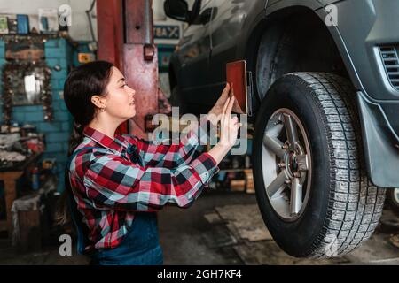 Portrait of a young Caucasian female mechanic, in uniform, using a gadget for modern car diagnostics. Side view. Indoors. Stock Photo