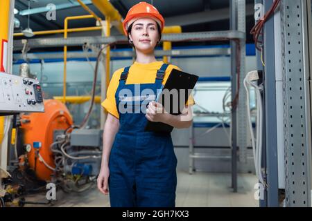 A female worker in helmet and uniform with a folder in her hand. In the background, boiler equipment and pipes. Concept of industrial production and f Stock Photo